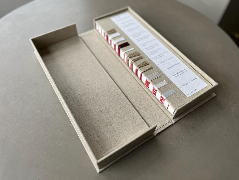 Op de afbeelding: Een gesloten, met linnen beklede doos met een rood-wit gestreept boek met stofstalen erin. Het boek bevat verschillende stofstalen en beschrijvingen.