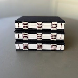 May include: Three black notebooks with white pages and burgundy stitching. The stitching is a geometric pattern.