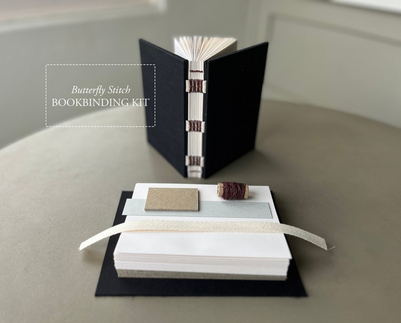 May include: A bookbinding kit with the text "Butterfly Stitch BOOKBINDING KIT" on a white background. The kit includes a black cover, a stack of white paper, a spool of brown thread, a piece of gray fabric, and a piece of brown cardboard.