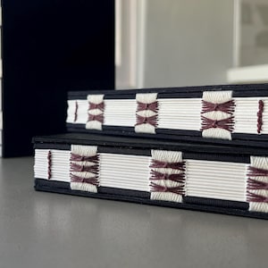 Op de afbeelding: Twee zwarte hardcover boeken met witte pagina's en decoratieve stiksels in bordeauxrood. De stiksels zijn in een geometrisch patroon.