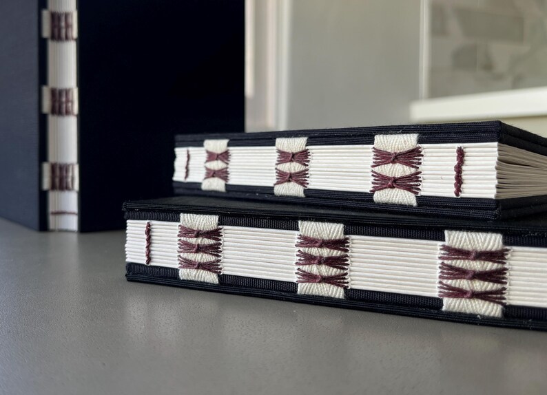 Op de afbeelding: Twee zwarte hardcover boeken met witte pagina's en decoratieve stiksels in bordeauxrood. De stiksels zijn in een geometrisch patroon.