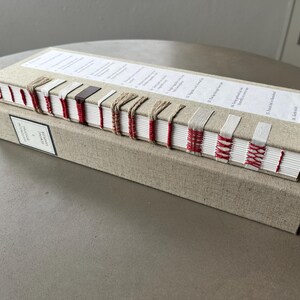 Op de afbeelding: Een in linnen gebonden boek met rode en witte stiksels op de rug. Het boek is getiteld "Een geschiedenis van de wereld in 100 objecten".