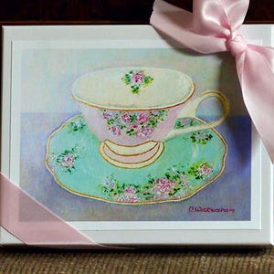 May include: A white teacup with pink floral designs sits on a saucer with a green and white floral pattern. The background is a soft purple and blue gradient. The image is signed "Wickesham".
