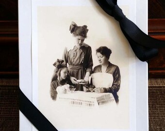 May include: A white gift box with a black and white photograph of three women and a child. The photograph is of a sepia tone. A black ribbon is tied around the box.