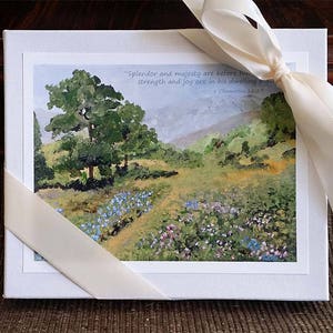 Könnte beinhalten: Eine weiße Schachtel mit einem Band darum gebunden. Die Schachtel zeigt ein Aquarellbild einer Landschaft mit Bäumen, Blumen und einem Zitat aus der Bibel: "Pracht und Hoheit sind vor ihm; Stärke und Freude sind in seiner Wohnung." 1 Chronik 16,27