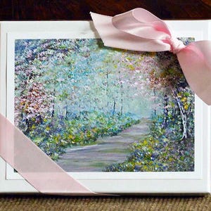 Puede incluir: Una caja blanca con una pintura de paisaje de un camino a través de un bosque, atada con un lazo de cinta rosa claro. La pintura tiene un estilo suave e impresionista con verdes, azules y toques de amarillo y rosa.