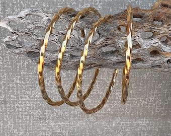 May include: Four gold-colored metal bangles with a twisted design. The bangles are stacked on top of each other and are displayed on a piece of driftwood.