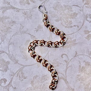 May include: A chain bracelet constructed from alternating silver-toned and copper-toned metal rings in a repeating pattern. The bracelet features a simple hook-style clasp at one end, with the links arranged in a curved, serpentine shape.