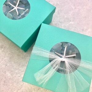 K&ouml;nnte beinhalten: Zwei t&uuml;rkisfarbene Geschenkboxen mit wei&szlig;em T&uuml;llband. Die Boxen haben ein rundes Fenster mit einem Seestern und Meerglas darin. Der Text "Blue Glass Design" ist auf der oberen Box aufgedruckt.