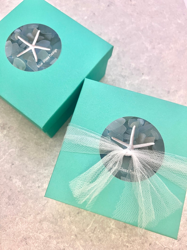 K&ouml;nnte beinhalten: Zwei t&uuml;rkisfarbene Geschenkboxen mit wei&szlig;em T&uuml;llband. Die Boxen haben ein rundes Fenster mit einem Seestern und Meerglas darin. Der Text "Blue Glass Design" ist auf der oberen Box aufgedruckt.
