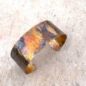 May include: A hammered brass cuff bracelet with a rustic, aged finish. The metal has a warm, golden hue with hints of copper and blue.
