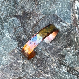 May include: A hammered copper bracelet with a rainbow iridescent finish. The bracelet is on a gray rock background.