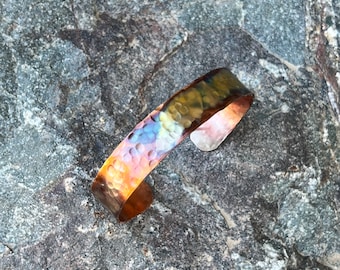 May include: A hammered copper bracelet with a rainbow iridescent finish. The bracelet is on a gray rock background.