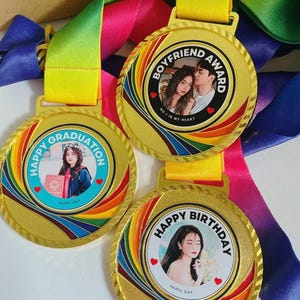 Puede incluir: Tres medallas doradas con detalles de arcoíris coloridos y cintas adjuntas. Cada medalla presenta una foto y el texto: "Happy Graduation", "Boyfriend Award" y "Happy Birthday". Las medallas tienen el texto "Happy Day" en la parte inferior.
