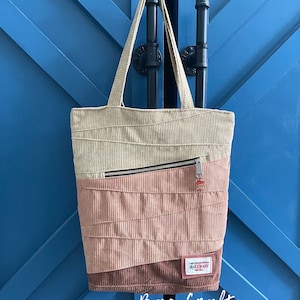 Może przedstawiać: Torba typu tote z beżowej i różowej tkaniny zamszowej z kieszenią zapinaną na zamek błyskawiczny i małą prostokątną etykietą z napisem "Designed & Handmade by BAE CRAFT".