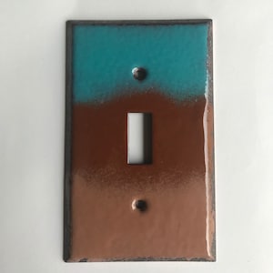Puede incluir: Una tapa de interruptor de luz con un diseño degradado azul turquesa y marrón. La tapa tiene una forma rectangular con esquinas redondeadas y una sola abertura para el interruptor.