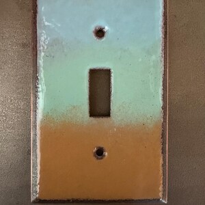 Puede incluir: Una placa de interruptor rectangular con un diseño degradado. La sección superior es azul claro, que pasa a verde y luego a un color marrón claro en la parte inferior. Tiene una abertura rectangular para el interruptor y dos orificios para tornillos.