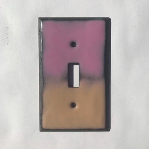Pode incluir: Uma capa decorativa para interruptor de luz com um gradiente de cor rosa a bege. O interruptor é branco com uma moldura preta, e a capa tem uma borda cinza escura. Design retangular.