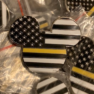 May include: A black and white Mickey Mouse pin with a thin yellow stripe across the middle. The Mickey Mouse head is shaped like the American flag with white stars on a black background. The bottom half of the Mickey Mouse head is shaped like a black and white striped flag with a thin yellow stripe across the middle.