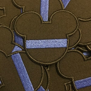 May include: Embroidered patches featuring a black Mickey Mouse head outline with a blue stripe. The patches are densely stitched with a black border and a blue stripe across the center of the head. The patches are made of a durable fabric.
