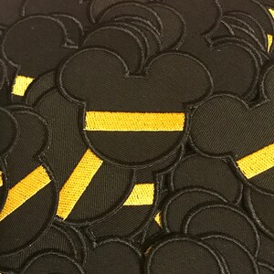 May include: A pile of black felt patches with a yellow stripe across the middle. The patches are shaped like a cartoon mouse head.