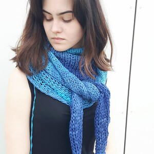 May include: A woman wearing a black dress with a blue and turquoise knitted scarf. The scarf is tied in a knot at the front and has a loose, open weave.