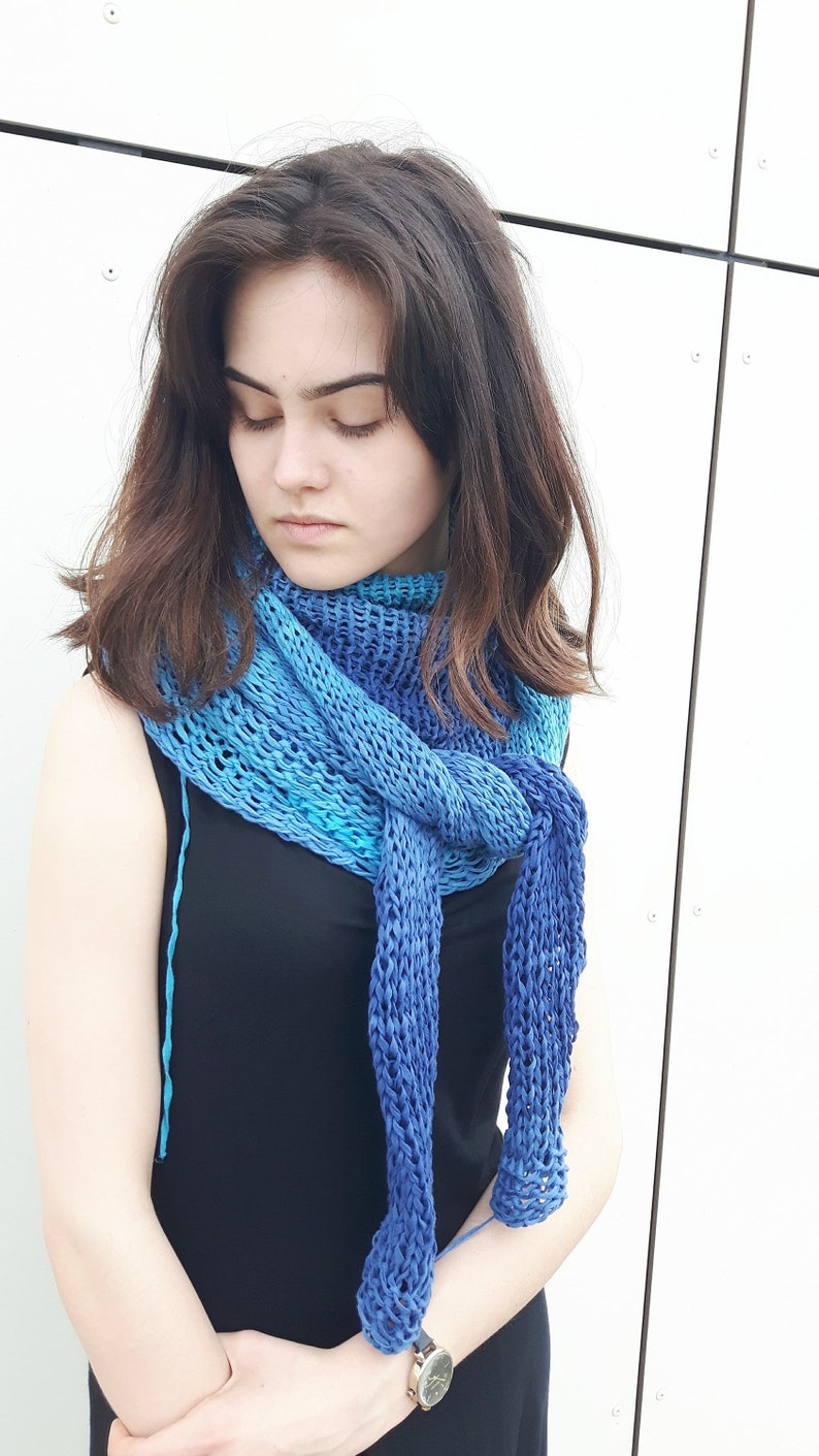 May include: A woman wearing a black dress with a blue and turquoise knitted scarf. The scarf is tied in a knot at the front and has a loose, open weave.