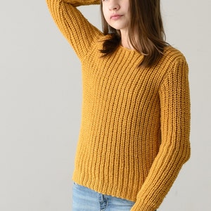 May include: A young person wearing a mustard yellow knitted sweater and blue jeans. The sweater has a ribbed texture.