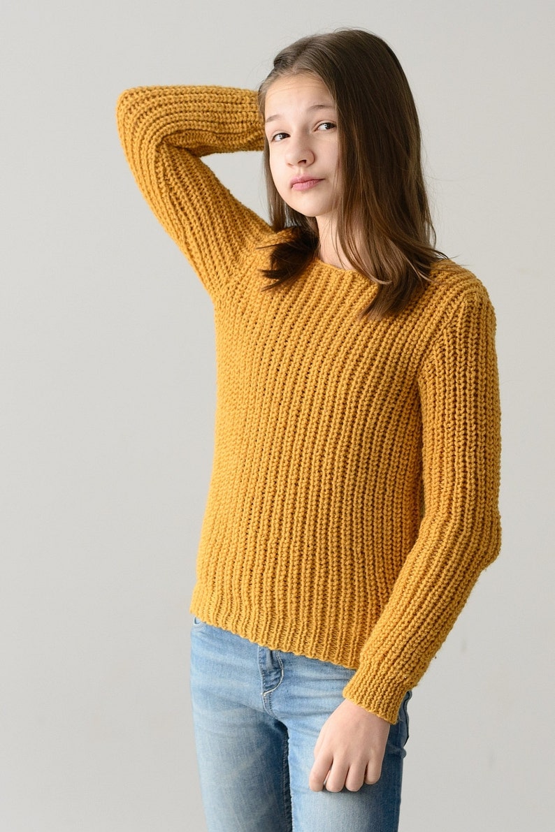 May include: A young person wearing a mustard yellow knitted sweater and blue jeans. The sweater has a ribbed texture.