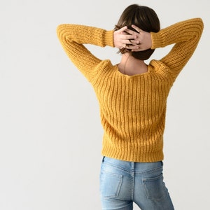 May include: A woman wearing a yellow knitted sweater and blue jeans. She is standing with her arms raised above her head.