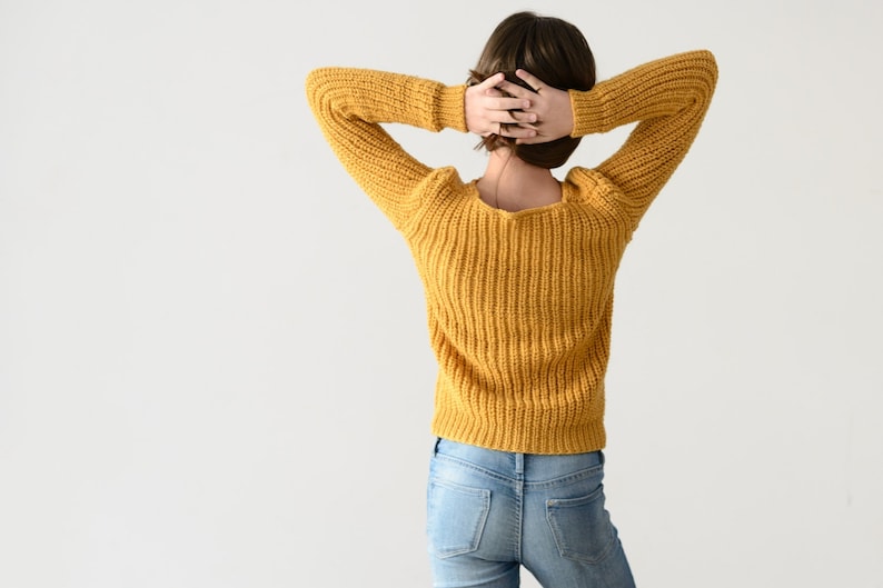May include: A woman wearing a yellow knitted sweater and blue jeans. She is standing with her arms raised above her head.