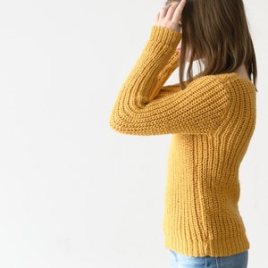 May include: A woman wearing a yellow knitted sweater and blue jeans. She is standing with her hand on her head.