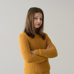May include: A young person with long brown hair wearing a yellow knitted sweater and blue jeans. They are standing with their arms crossed in front of a white background.
