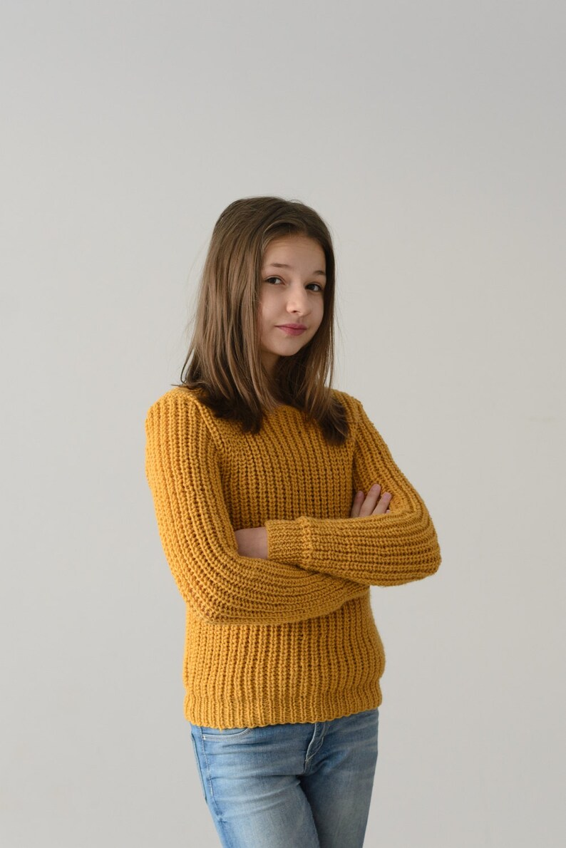 May include: A young person with long brown hair wearing a yellow knitted sweater and blue jeans. They are standing with their arms crossed in front of a white background.