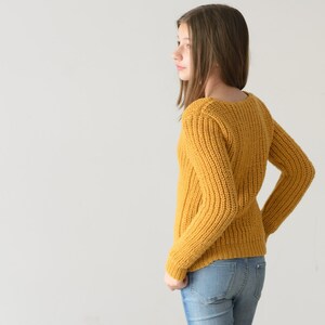 May include: A young person wearing a yellow knitted sweater and blue jeans. The sweater has a ribbed texture and a scoop neck.
