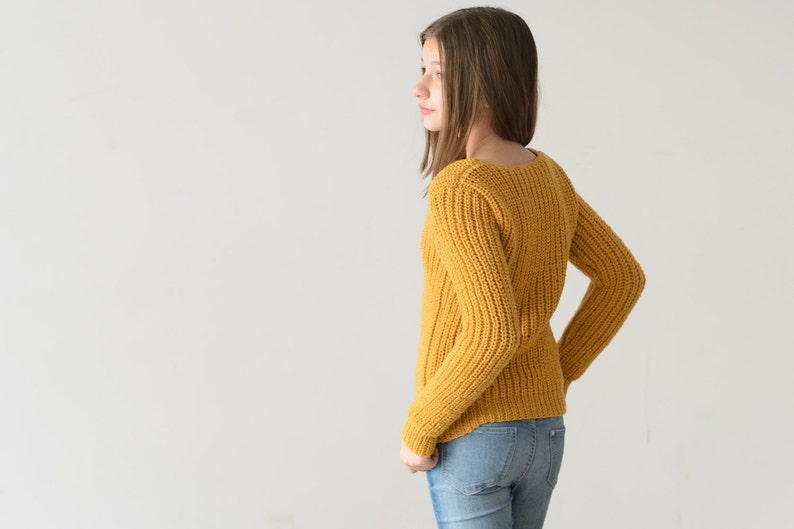 May include: A young person wearing a yellow knitted sweater and blue jeans. The sweater has a ribbed texture and a scoop neck.
