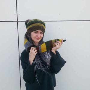 Könnte beinhalten: Eine Frau trägt eine grün-gelb-braun gestreifte Strickmütze mit passendem Schal. Die Mütze hat eine lange, spitze Spitze, die am Hinterkopf herunterhängt.