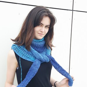 May include: A woman wearing a black dress with a blue and teal knitted scarf. The scarf is tied around her neck and draped over her shoulders.
