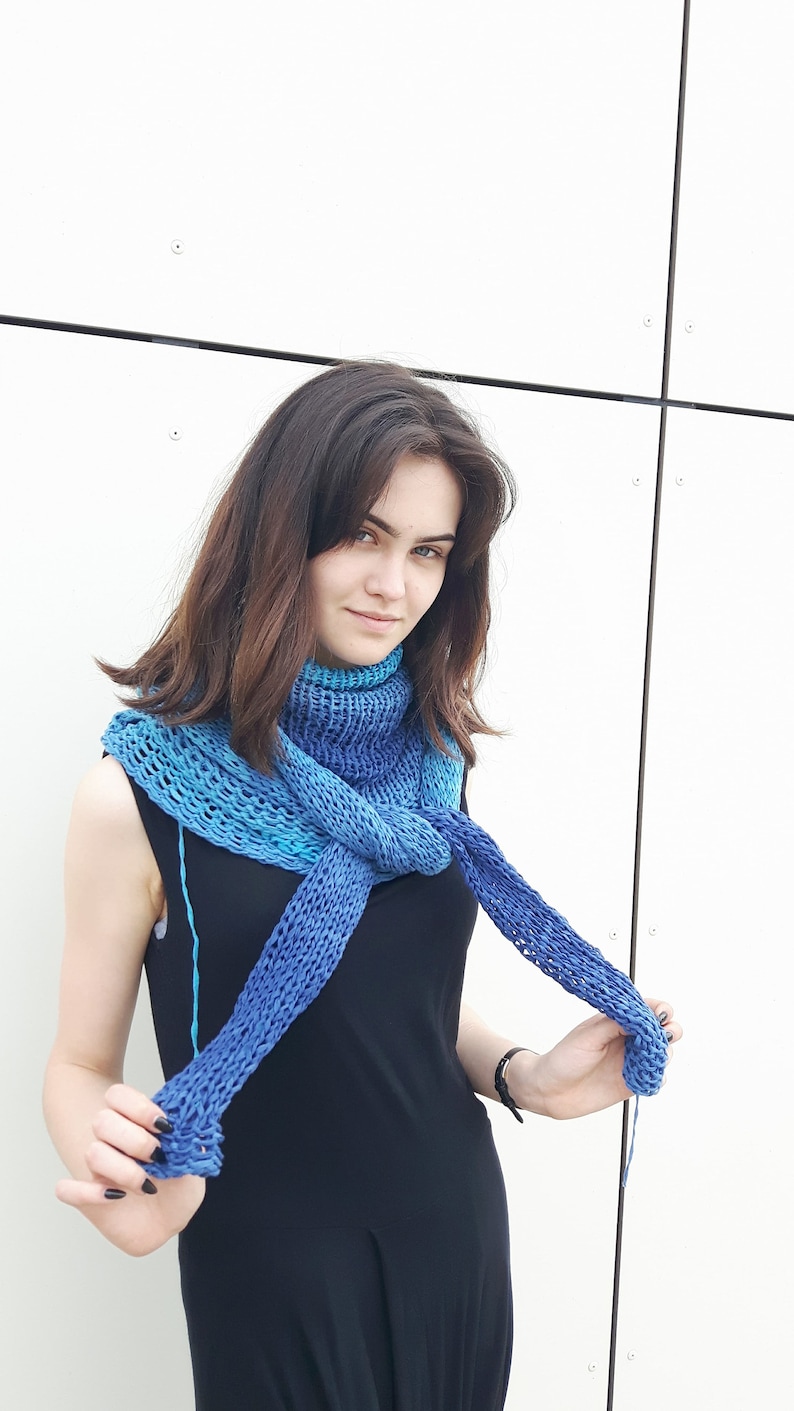 May include: A woman wearing a black dress with a blue and teal knitted scarf. The scarf is tied around her neck and draped over her shoulders.