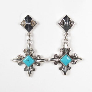 May include: A pair of silver dangle earrings with turquoise stones set in a geometric design. The earrings feature a square top with a smaller square in the center and a dangling cross-shaped design with a turquoise stone in the center.