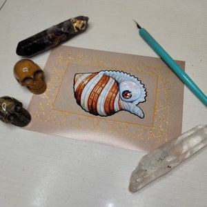 May include: A detailed illustration of a seashell with brown and white stripes, an eye, and a red ladybug. The artwork is on a pinkish-brown card with a gold border. Additional crystals and skull figurines surround the card.