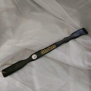 May include: A long, black wooden shoehorn featuring the gold-colored text Steelers and a small circular Steelers team logo emblem. The handle end includes a small hole with a thin brown twine loop attached for hanging the item.