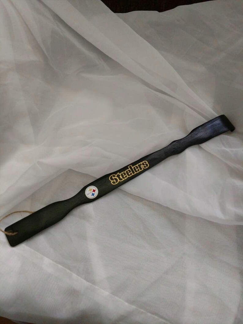 May include: A long, black wooden shoehorn featuring the gold-colored text Steelers and a small circular Steelers team logo emblem. The handle end includes a small hole with a thin brown twine loop attached for hanging the item.