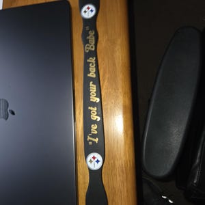 May include: A long, narrow, black wooden paddle featuring the gold text "I've got your back Babe" centered on the handle. Two circular Pittsburgh Steelers team logos are positioned above and below the text on the flat surface of the paddle.
