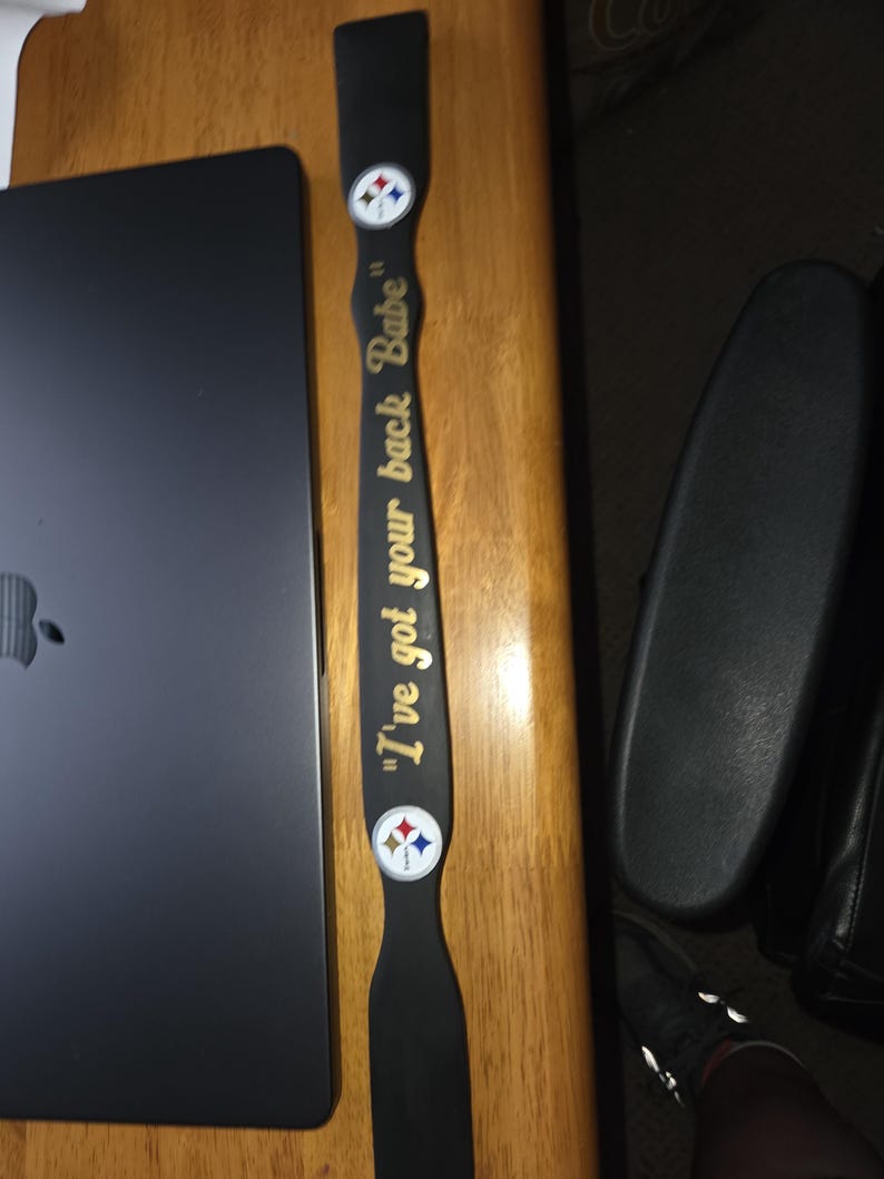 May include: A long, narrow, black wooden paddle featuring the gold text "I've got your back Babe" centered on the handle. Two circular Pittsburgh Steelers team logos are positioned above and below the text on the flat surface of the paddle.