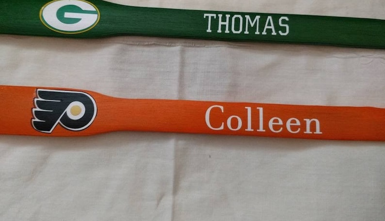 May include: Two wooden paddles featuring sports team logos and names. The top green paddle displays a white G logo and the name THOMAS in white block letters. The bottom orange paddle features a black, white, and orange logo with the name Colleen.