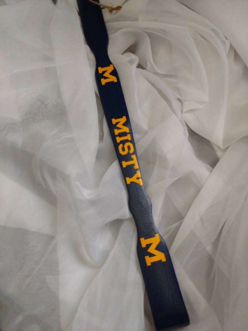 May include: A long, dark blue wooden paddle with a smooth, contoured handle. The word MISTY is printed in bold, yellow, block-style capital letters vertically down the center, flanked by a yellow letter M at the top and bottom of the text.