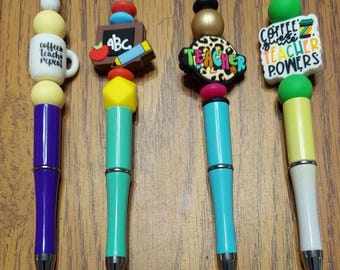 May include: Four colorful pens with different charms. The charms include a coffee cup with the text "coffee teach repeat", a chalkboard with the letters "ABC", a leopard print charm with the word "teacher", and a green charm with the text "coffee gives teacher powers".