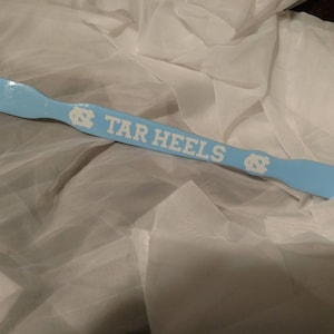 May include: A light blue shoehorn featuring the white text TAR HEELS and two white North Carolina Tar Heels logos. The shoehorn has a smooth, glossy finish and a curved handle end with a small hole for hanging, displaying a sports team theme.