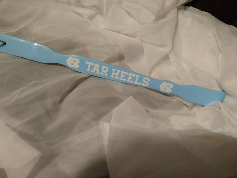 May include: A light blue shoehorn featuring the white text TAR HEELS and two white North Carolina Tar Heels logos. The shoehorn has a smooth, glossy finish and a curved handle end with a small hole for hanging, displaying a sports team theme.